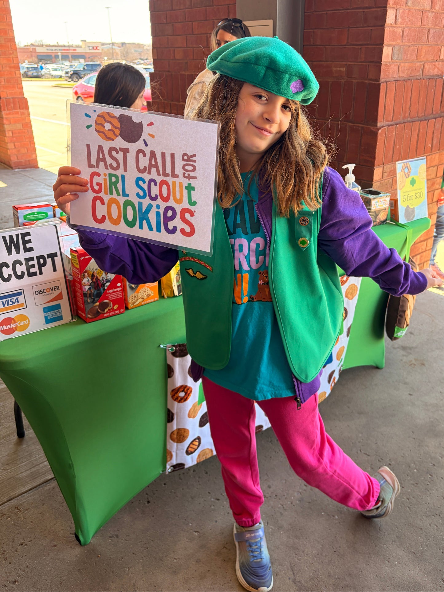 Young girl in Girl Scout uniform holding a sign with text, standing on a green chair outdoors.