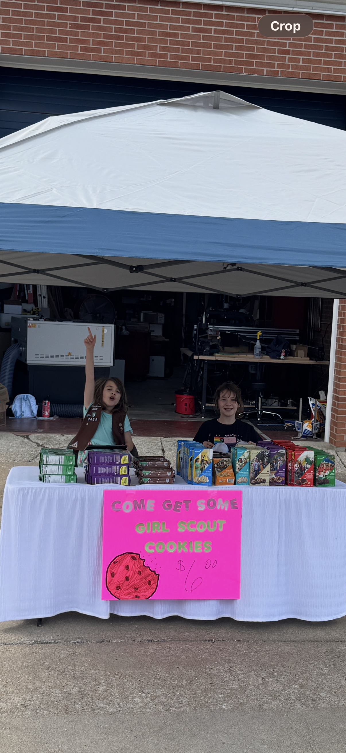 Two people behind a table with  cookies under a tent, with a pink sign reading 'Come Get Some Girl Scout Cookies $6' 